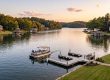 Pontoon boat with people on a calm lake at sunset, with shoreline homes and a wooden dock nearby.