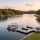 Pontoon boat with people on a calm lake at sunset, with shoreline homes and a wooden dock nearby.
