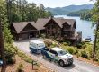 Lakeside log cabin with a gravel driveway, pines, and a white pickup pulling a Property Stewards trailer in front of the house.