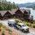 Lakeside log cabin with a gravel driveway, pines, and a white pickup pulling a Property Stewards trailer in front of the house.