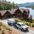 Lakeside log cabin with a gravel driveway, pines, and a white pickup pulling a Property Stewards trailer in front of the house.