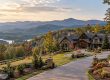 Luxury stone-and-wood house perched on a hillside with a curved driveway and autumn-colored trees, overlooking distant mountains.