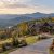 Luxury stone-and-wood house perched on a hillside with a curved driveway and autumn-colored trees, overlooking distant mountains.