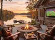 Lakeside patio at sunset with a stone fire pit, four wooden chairs draped in plaid blankets, and a TV showing a football game on the house wall overlooking a docked lake.