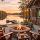 Lakeside patio at sunset with a stone fire pit, four wooden chairs draped in plaid blankets, and a TV showing a football game on the house wall overlooking a docked lake.