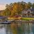 Lakeside luxury home with a dock, boat, and autumn trees along the shore.