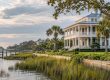 White two-story coastal house with a wraparound porch along a marshy waterfront, palm trees, and a wooden dock extending into calm water.