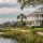 White two-story coastal house with a wraparound porch along a marshy waterfront, palm trees, and a wooden dock extending into calm water.
