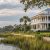 White two-story coastal house with a wraparound porch along a marshy waterfront, palm trees, and a wooden dock extending into calm water.