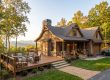 Cozy wooden mountain cabin with a large deck and stone columns, autumn trees surround the house, and a car in the driveway.